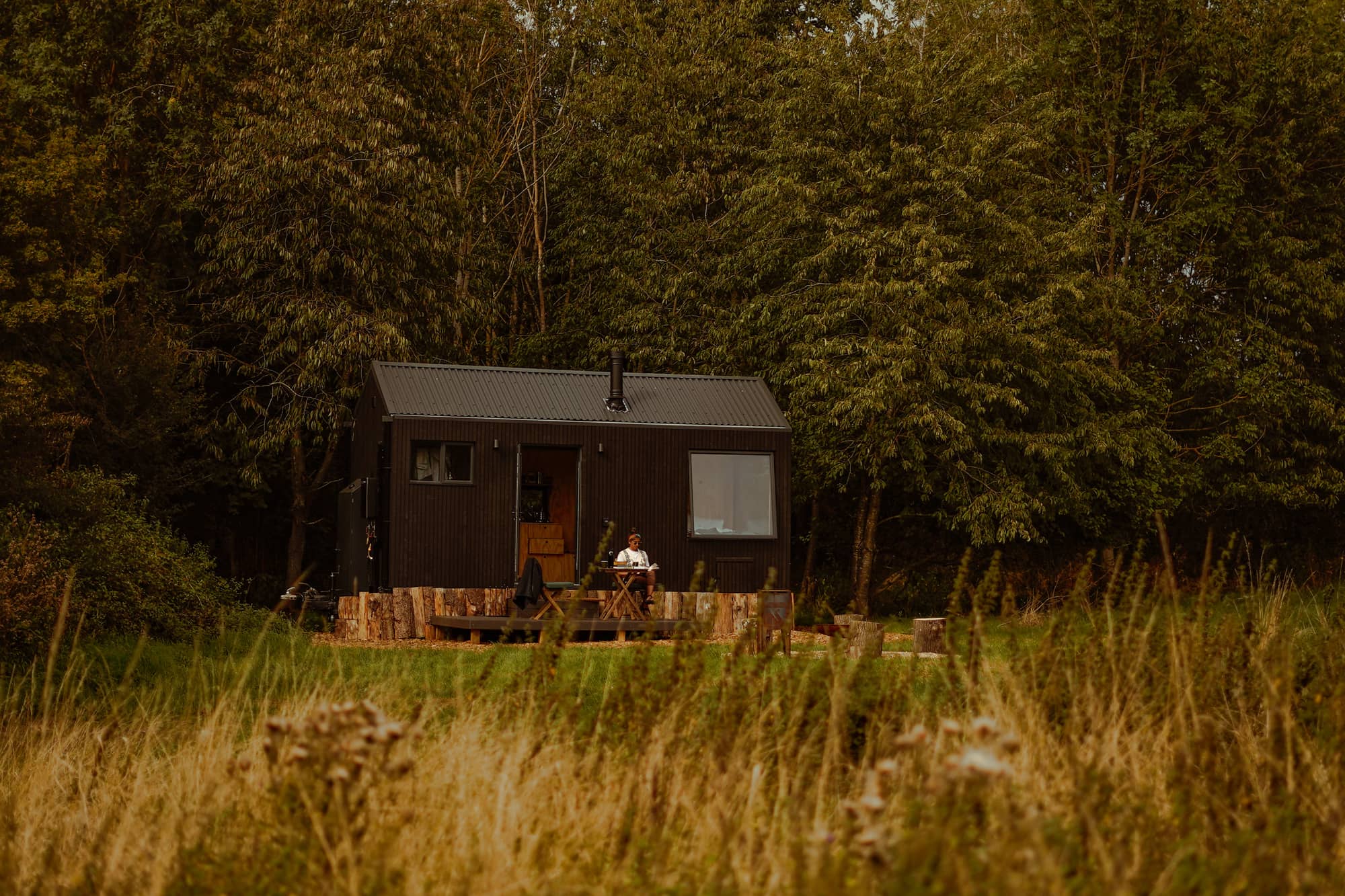 An Unyoked cabin in a forest clearing, someone reading at a table outside.
